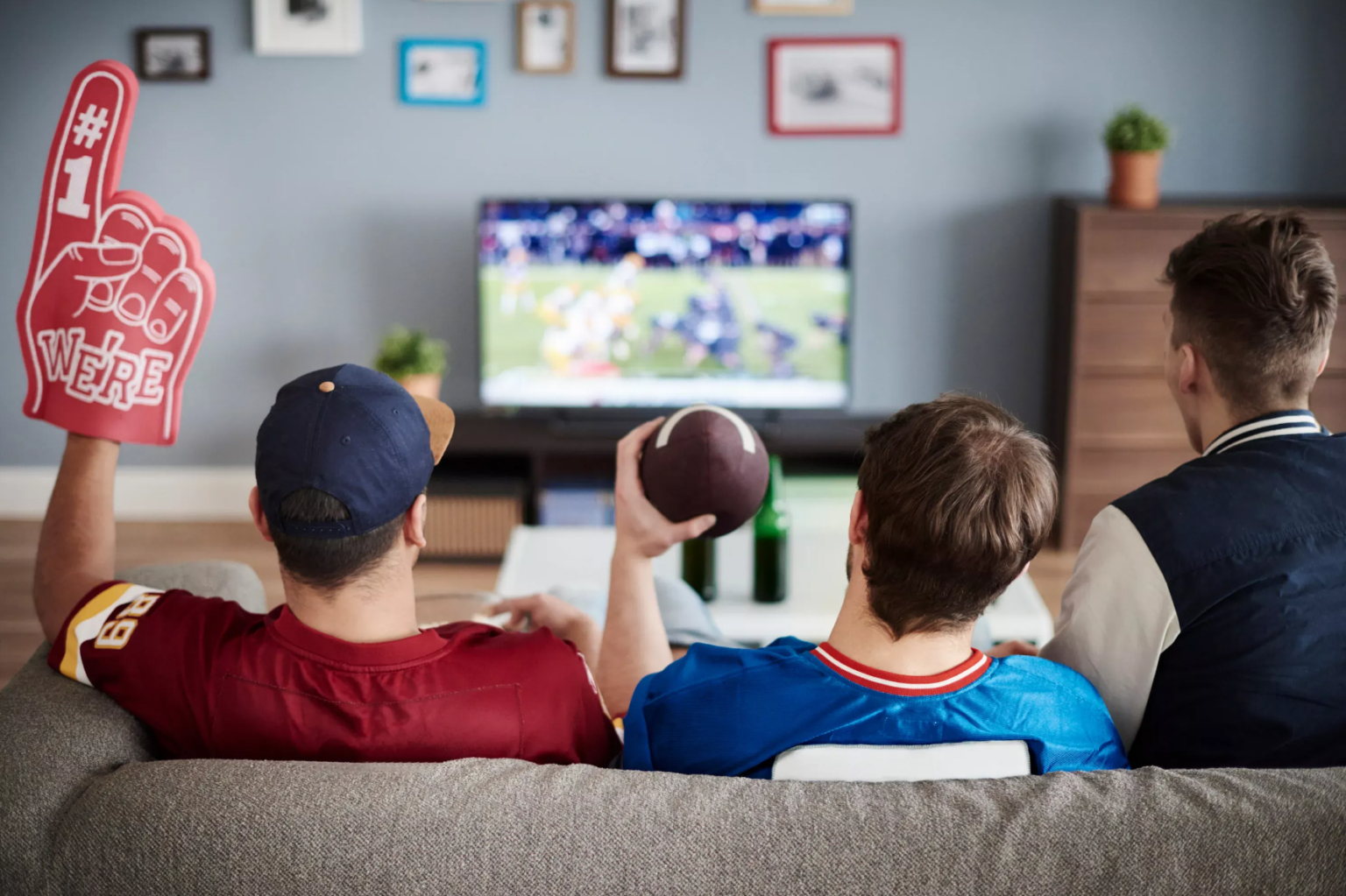 Three guys sitting on a couch watching football, holding a foam finger and a football